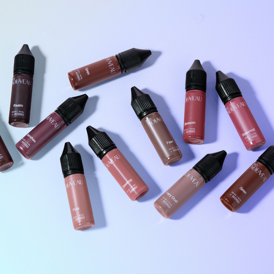 Top-down view of several Pro Nouveau pigment bottles in various berry, mauve, and nude shades scattered on a light purple gradient background.